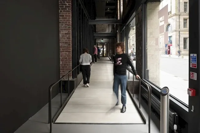 A ramp between exhibition spaces in the Fruitmarket Gallery — wide, shallow, with handrails on both sides, with visitors using it naturally